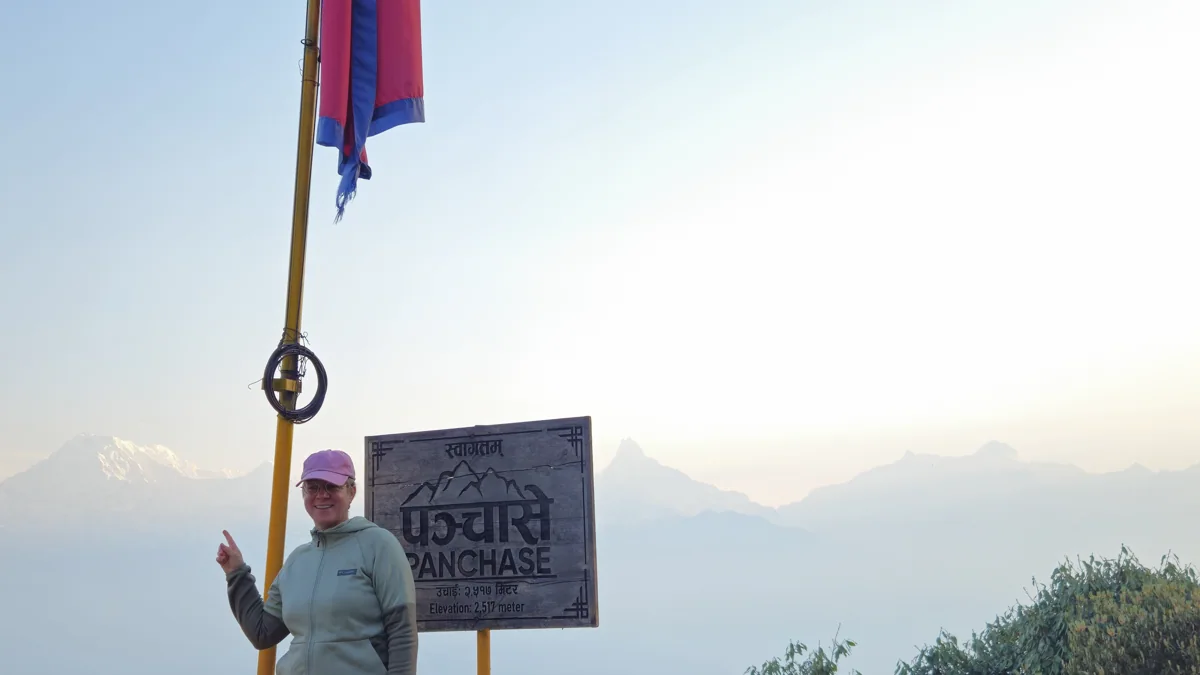 A trekker beside the Panchase summit sign and Nepal flag with layered hills behind